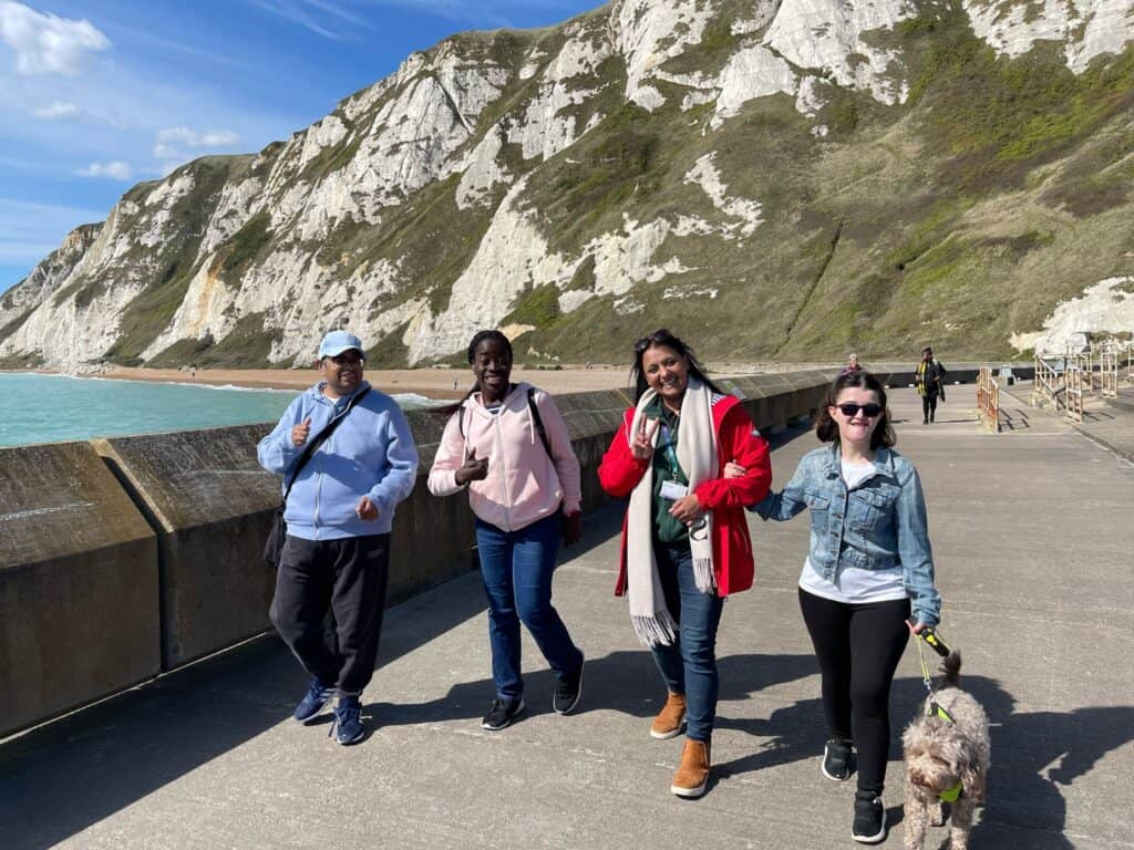 Four people and a dog walk along a coastal path with white cliffs and blue sea in the background.