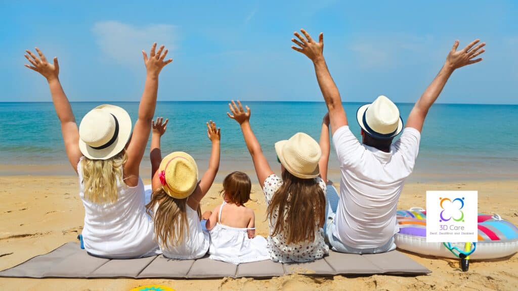 Five people sitting on a beach towel facing the ocean, all raising their arms, with hats on. A float and a logo are on the sand nearby.