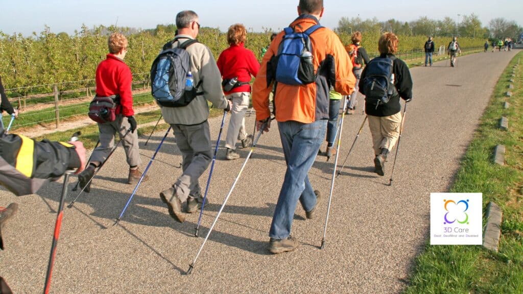 A group of people with backpacks walk on a paved path using Nordic walking poles; trees line the road, and a logo reads "3D Care.