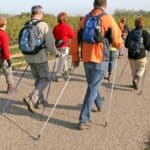 A group of people with backpacks walk on a paved path using Nordic walking poles; trees line the road, and a logo reads "3D Care.